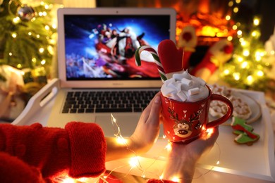 Photo of MYKOLAIV, UKRAINE - DECEMBER 23, 2020: Woman with sweet drink watching The Christmas Chronicles movie on laptop near fireplace at home, closeup. Cozy winter holidays atmosphere