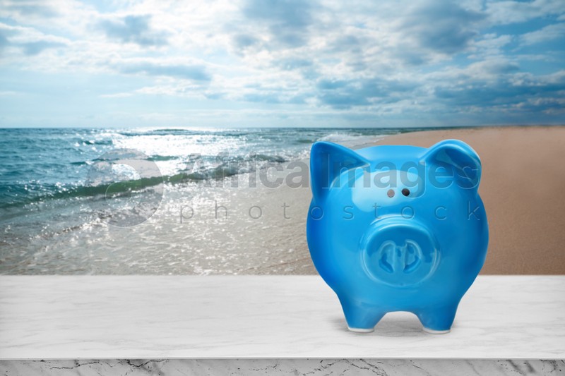 Saving money for summer vacation. Piggy bank on white marble surface near sandy beach and sea, space for text