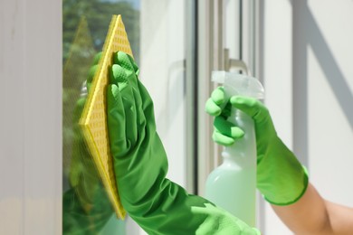 Woman cleaning window glass with sponge cloth and spray indoors, closeup