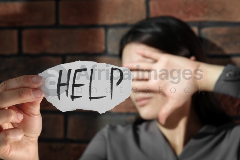 Photo of Domestic violence concept. Unhappy woman holding paper with written word Help near brick wall, focus on hand