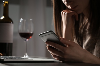 Woman using smartphone at table with laptop near glass of wine indoors, closeup. Loneliness concept