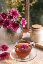 Beautiful chrysanthemum flowers in vase and cup of tea on beige textured table near window. Autumn still life