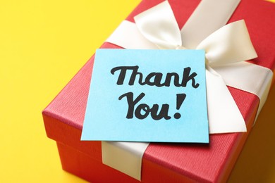 Gift box and light blue paper note with phrase Thank You on yellow background, closeup