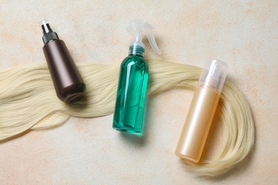Photo of Spray bottles with thermal protection and lock of blonde hair on beige textured table, flat lay