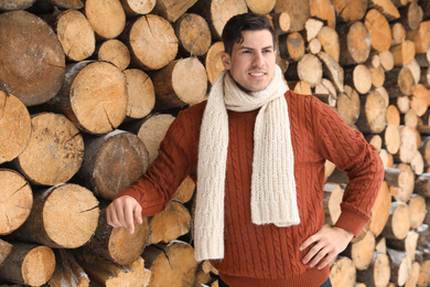Handsome man wearing warm sweater and scarf near stack of firewood. Winter season
