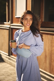 Beautiful young woman in stylish blue dress with handbag and cup of coffee near wooden building outdoors