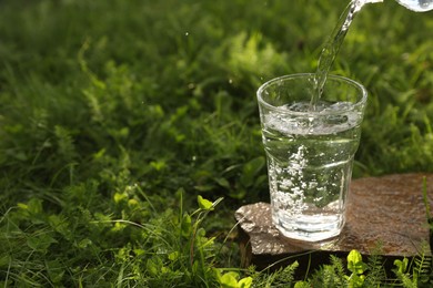 Photo of Pouring fresh water from bottle into glass on stone in green grass outdoors. Space for text