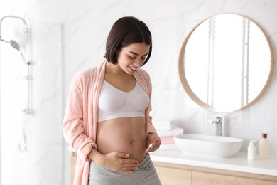 Young pregnant woman applying cosmetic product on belly in bathroom