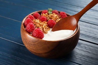 Eating tasty yogurt with raspberries and muesli from bowl on blue wooden table