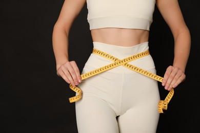 Woman in sportswear measuring waist with tape on black background, closeup