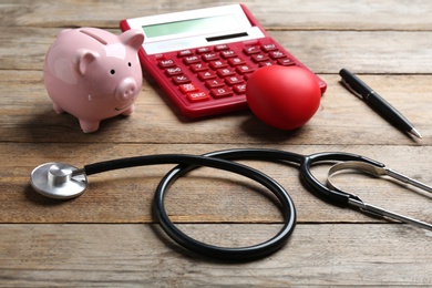 Stethoscope, red heart and calculator with piggybank on wooden surface. Health insurance concept