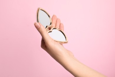 Woman holding stylish heart shaped cosmetic pocket mirror on pink background, closeup