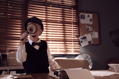 Cute little detective with magnifying glass at table in office. Space for text