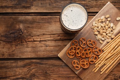 Glass of beer served with delicious pretzel crackers and other snacks on wooden table, flat lay. Space for text