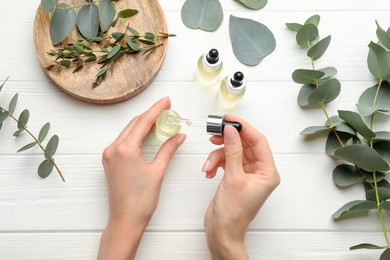 Woman taking eucalyptus essential oil with dropper from bottle at white wooden table, top view