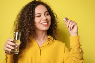 African-American woman with glass of water and vitamin pill on yellow background