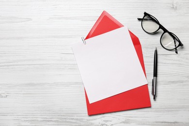 Envelope with blank paper card, glasses and pen on white wooden table, flat lay. Space for text
