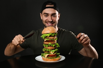 Photo of Young man with cutlery eating huge burger on black background
