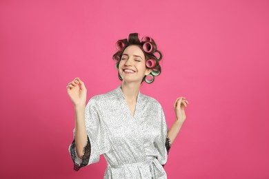Happy young woman in silk bathrobe with hair curlers dancing on pink background
