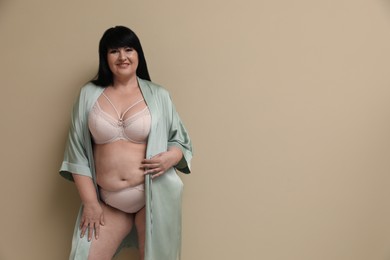 Beautiful overweight woman in sexy underwear and silk robe on beige background, space for text. Plus-size model