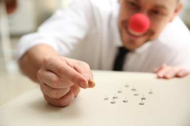 Man with clown nose putting pins onto colleague's chair in office, focus on hand. Funny joke