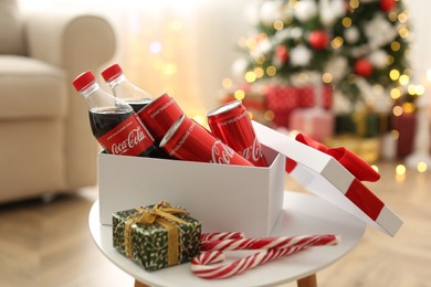Photo of MYKOLAIV, UKRAINE - JANUARY 15, 2021: Gift box with Coca-Cola cans and bottles on table in room decorated for Christmas