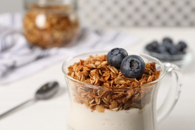 Photo of Tasty yogurt with muesli and blueberries in cup served on white table, closeup
