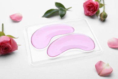 Photo of Package with under eye patches and rose flowers on white background, closeup. Cosmetic product