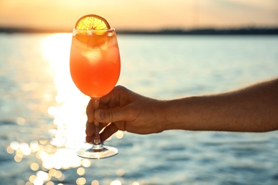 Man with glass of fresh summer cocktail near water at sunset, closeup