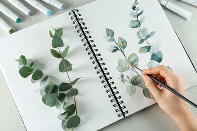 Woman drawing beautiful eucalyptus branches in sketchbook at white table, top view