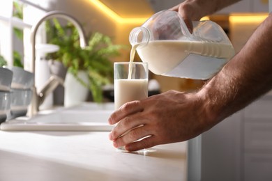 Man pouring milk from gallon bottle into glass at white countertop in kitchen, closeup