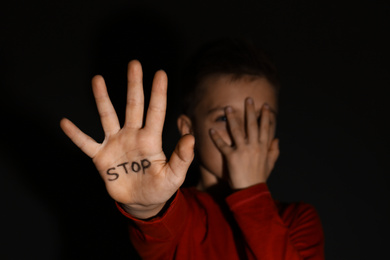 Photo of Abused little boy showing palm with word STOP against black background, focus on hand. Domestic violence concept