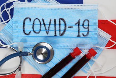 COVID-19 written on protective mask, test tubes with blood, stethoscope and American flag, flat lay. Coronavirus pandemic in USA