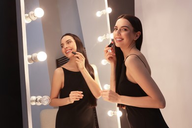 Photo of Beautiful young woman in elegant dress with lipstick near mirror indoors