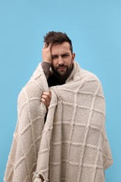 Man wrapped in blanket suffering from headache on light blue background. Cold symptoms