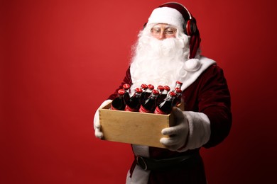 MYKOLAIV, UKRAINE - JANUARY 18, 2021: Santa Claus with headphones holding wooden crate full of Coca-Cola bottles on red background