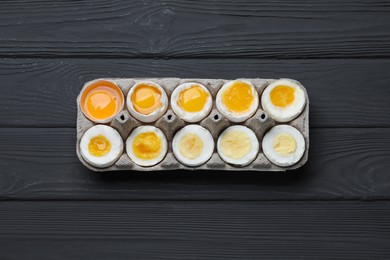 Photo of Boiled chicken eggs of different readiness stages in carton on black wooden table, top view