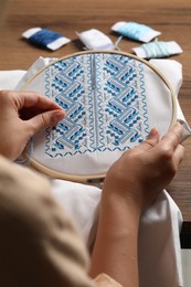 Photo of Woman embroidering white shirt with blue thread at wooden table, closeup. Ukrainian national clothes