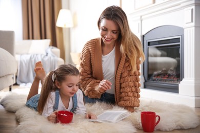 Happy woman and her daughter reading book near fireplace at home