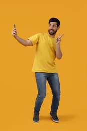Photo of Smiling man taking selfie with smartphone and showing peace sign on yellow background