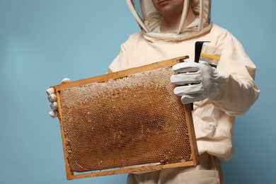 Beekeeper in uniform holding hive frame with honeycomb and tools on light blue background, closeup