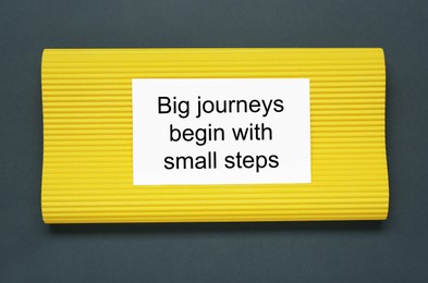 Photo of Card with phrase Big Journeys Begin With Small Steps on dark background, top view. Motivational quote