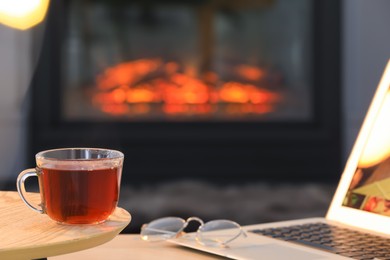 Glass cup of hot tea on wooden table near fireplace at home, space for text. Cozy atmosphere