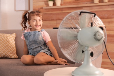 Little girl enjoying air flow from fan on sofa in living room. Summer heat