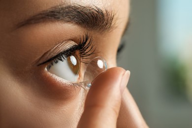 Young woman putting contact lens in her eye on blurred background, closeup