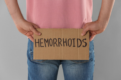 Man holding carton sign with word HEMORRHOIDS on light grey background, closeup