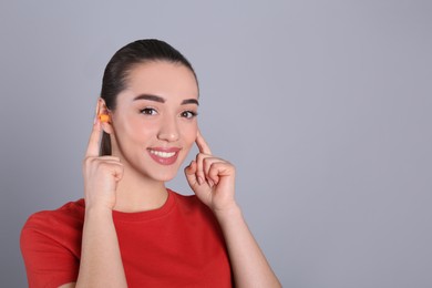 Young woman inserting foam ear plugs on grey background. Space for text
