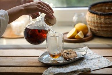 Photo of Woman pouring delicious tea into glass at wooden table, closeup