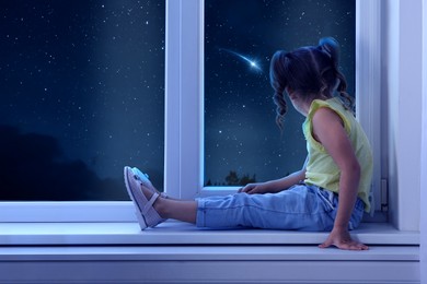 Image of Cute little girl sitting near window and looking at shooting star in beautiful night sky
