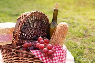 Wicker picnic basket with bottle of wine, bread, grapes and napkin on green grass outdoors, closeup. Space for text